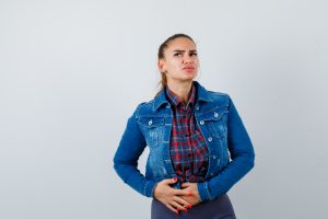 Young woman with hands on stomach in checkered shirt, jean jacket and looking painful , front view.